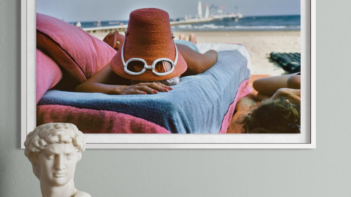 A Vintage beach photograph by Slim Aarons of a woman tanning with a hat and a pair of white glasses