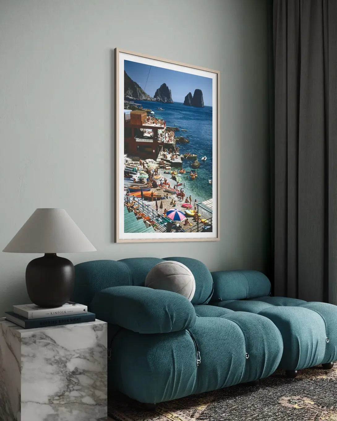 A framed vertical art print of a colorful coastal village and sea scene, displayed on a sage green wall above a teal modular sofa in a modern living room setting.