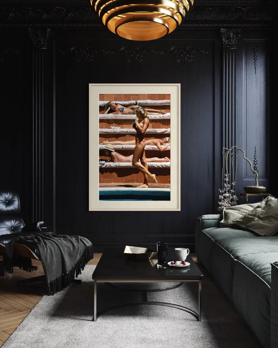 A framed art print featuring a vintage-style photograph of a woman in a bikini lounging on stacked wooden lounge beds, displayed on a dark paneled wall in a modern, moody living room setting.