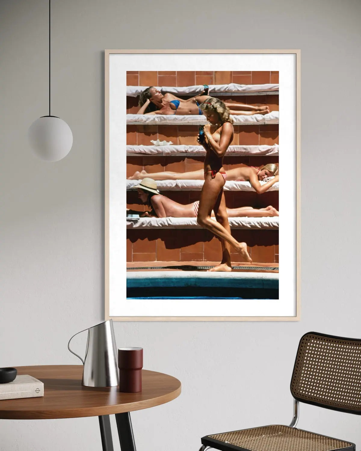 A framed art print featuring a photograph of women relaxing by a swimming pool, hung on a light gray wall in a modern room setting with a wooden table and chair.