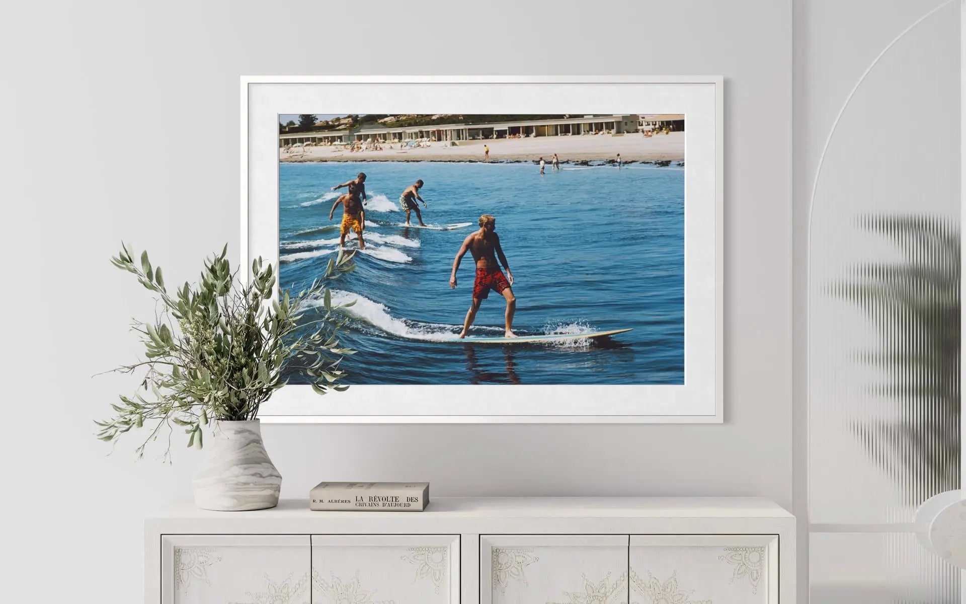 A framed photographic print of surfers riding waves at a beach, displayed on a white wall above a white cabinet with a vase of greenery.
