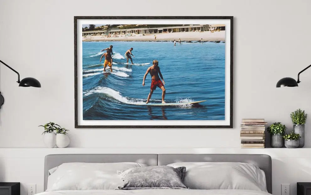 A framed wall art print depicting surfers riding blue ocean waves on a beach, displayed above a bed in a modern bedroom setting with black wall lamps and small plants.