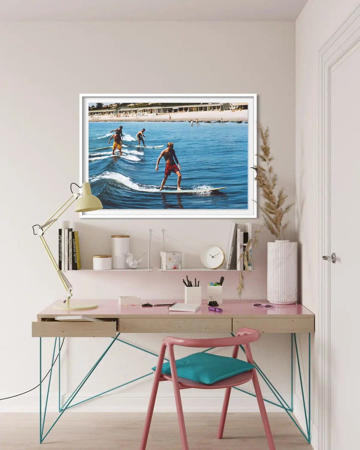 A framed art print depicting three surfers riding waves in a sunny beach setting, displayed on a light pink desk shelf in a modern, styled home office environment with a desk, chair, and lamp.