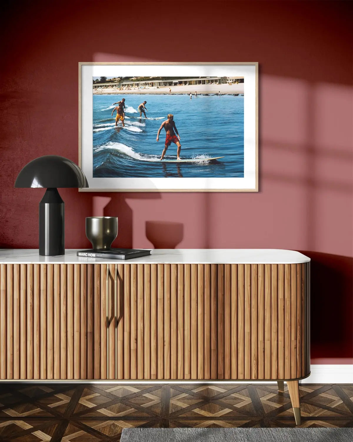 A framed art print depicting surfers riding blue ocean waves, displayed on a reddish-brown wall above a modern wooden console table with a black lamp.