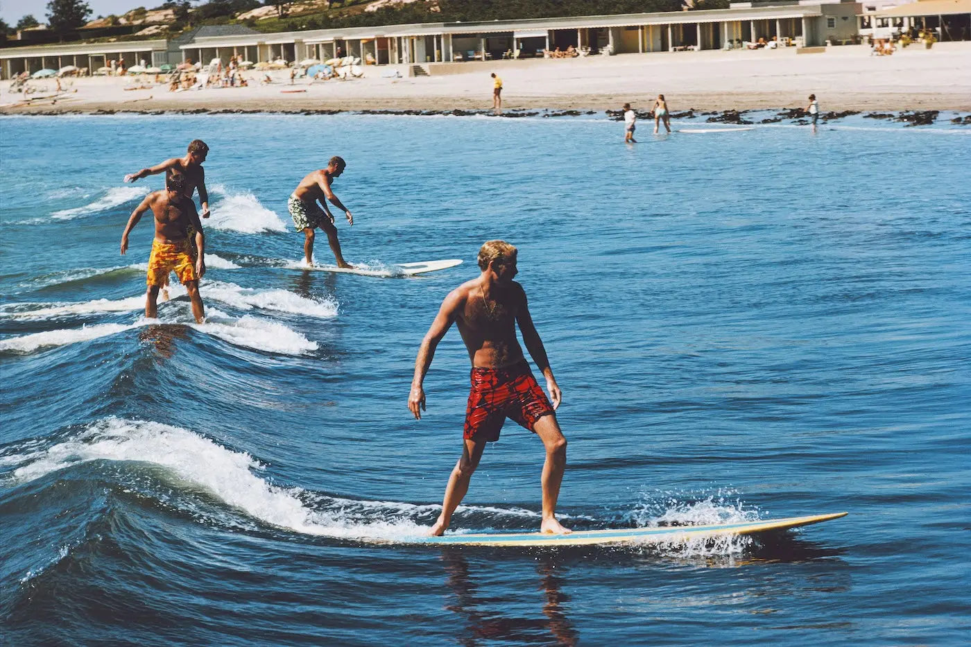 A man in red patterned swim trunks stands on a surfboard in blue ocean water, with other surfers and a beach with buildings in the background.