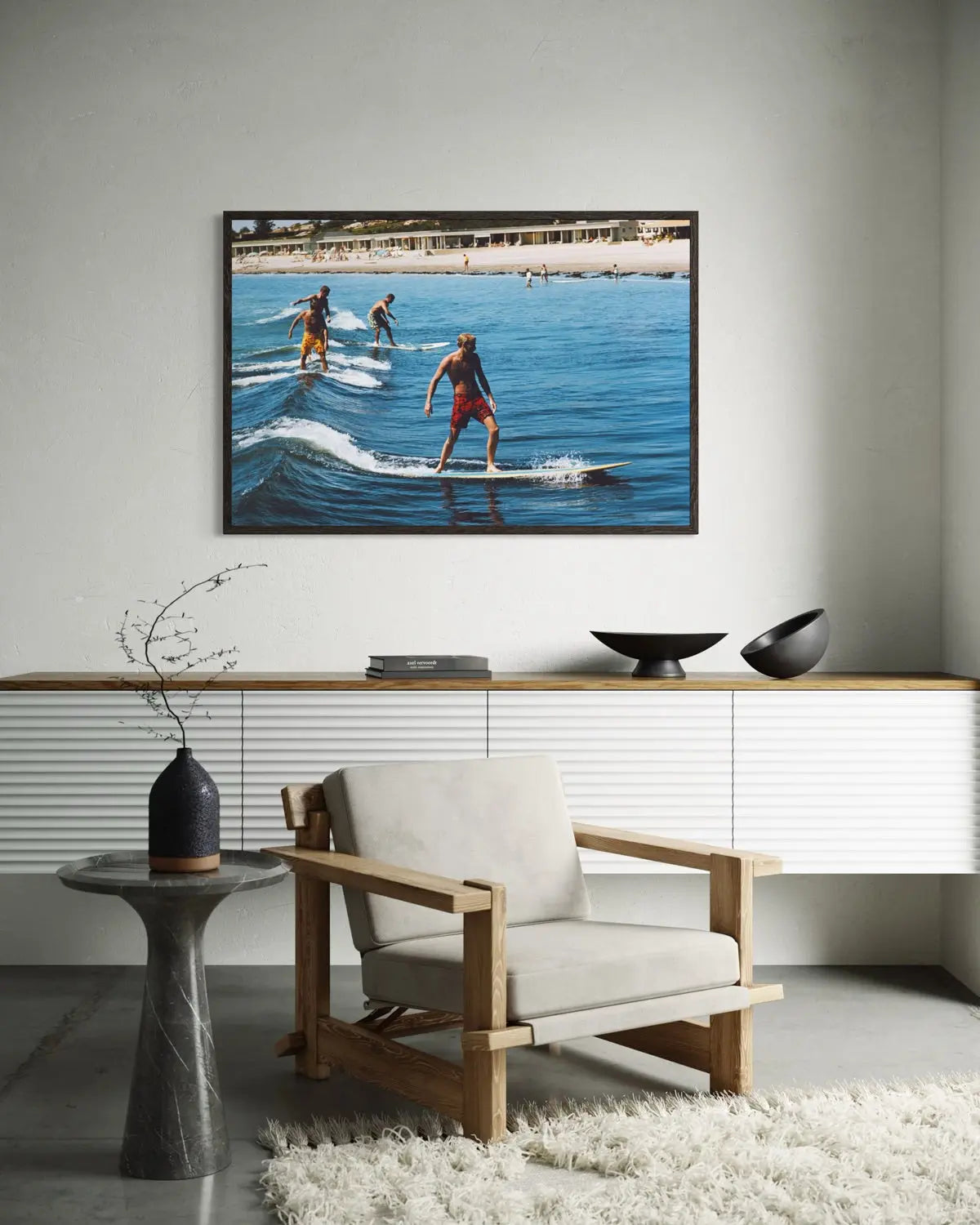 A framed square wall art print showing people surfing on blue ocean water, displayed above a white sideboard in a modern living room with a wooden armchair, side table, and white shag rug.