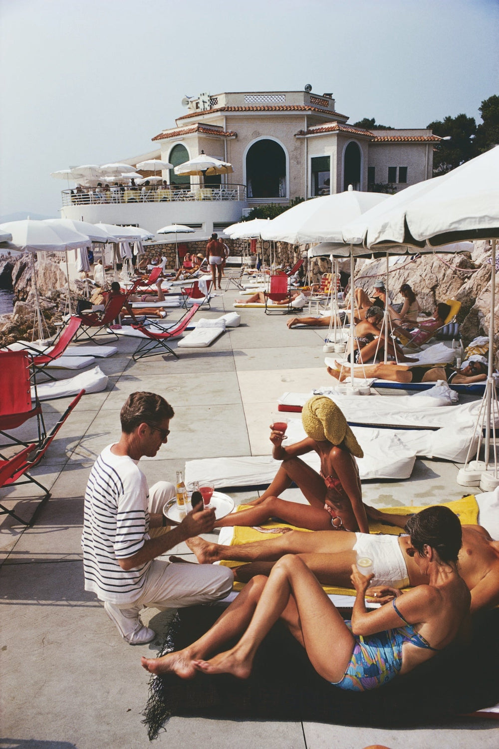 A sunlit resort poolside scene with people lounging on white lounge chairs under large umbrellas, a waiter serving drinks, and a large white building in the background.