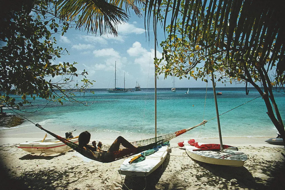 Person relaxing in a hammock on a tropical beach with boats and palm trees.