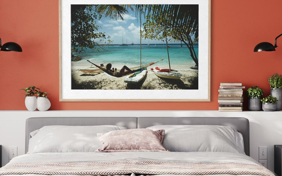 A framed beach scene print hangs on a terracotta-colored wall above a bed. The artwork shows palm trees, a sandy shore, turquoise water, and small boats, while the room includes a gray headboard, white bedding, black wall lamps, and small plants.