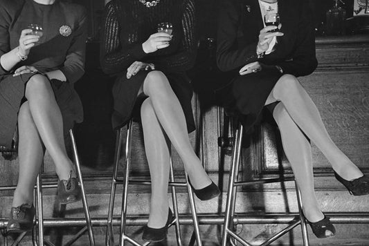 A black and white photo featuring three pairs of stockinged legs seated on stools, a vintage photograph.
