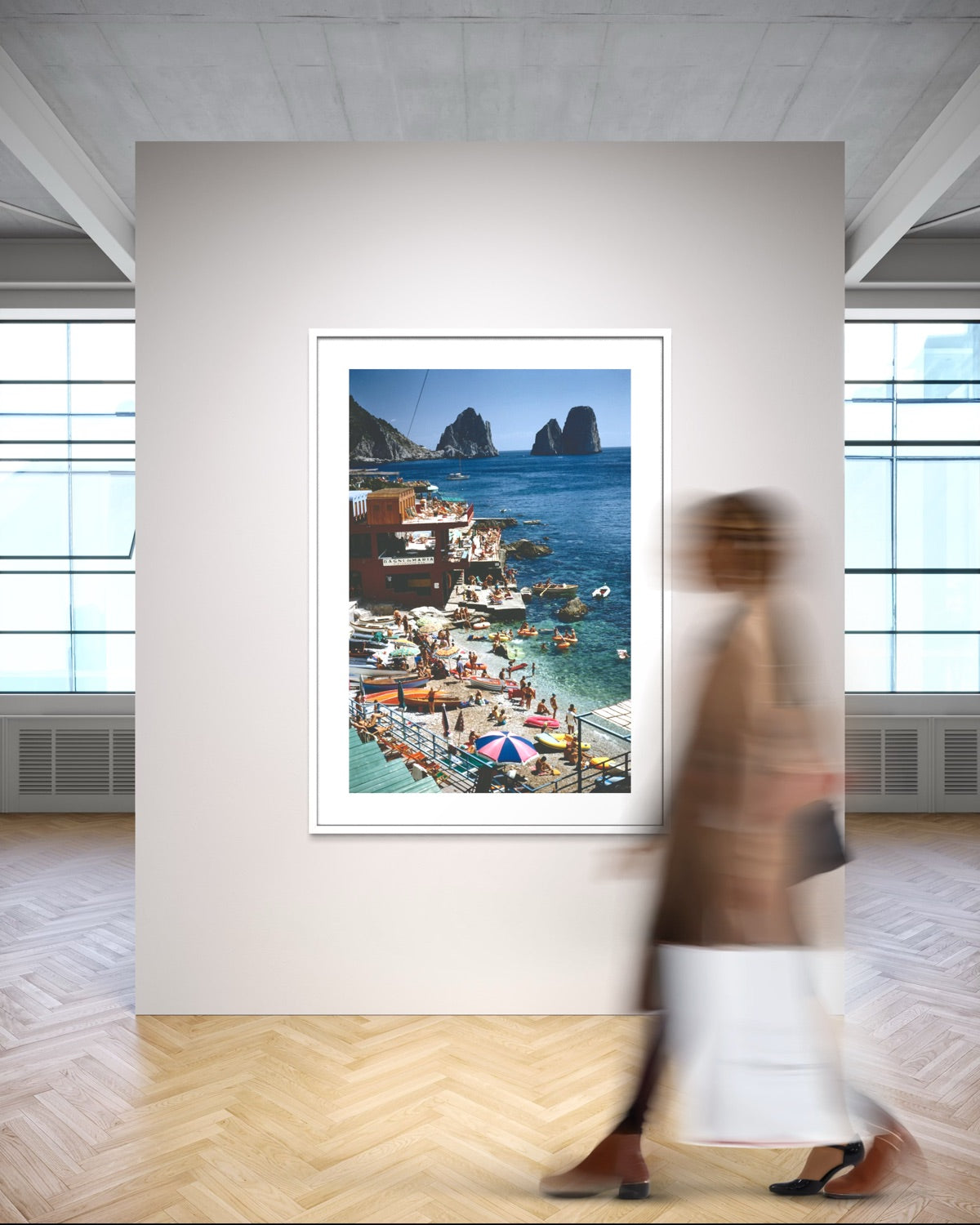 A framed coastal art print depicting a colorful seaside village with boats and rocky cliffs, displayed on a wall in a modern gallery space with a blurred person walking past in the foreground.