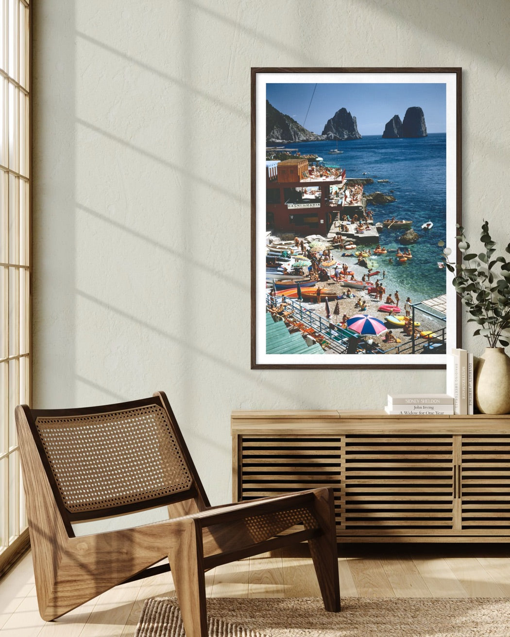 A framed vertical art print depicting a vibrant coastal harbor scene with boats, colorful umbrellas, and rocky cliffs, styled in a modern interior next to a wooden chair and console table.