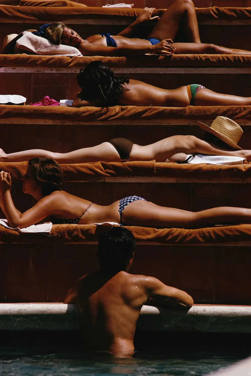 People sunbathing on steps by a poolside with a person in the water. The Picturalist Slim Aarons Collection.