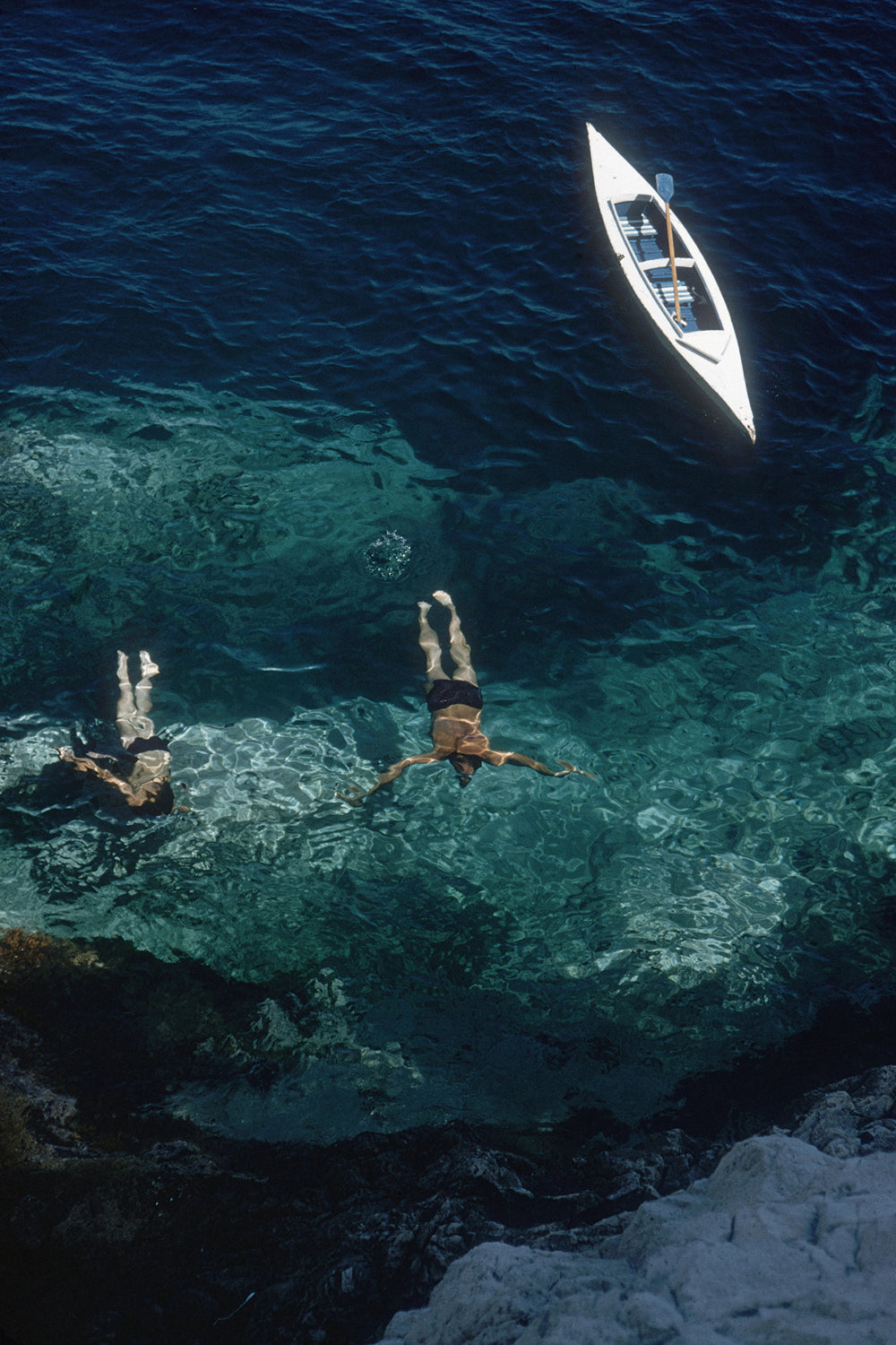 Aerial view of two people swimming in clear turquoise water near a small white boat on deep blue water, with rocky shoreline in the foreground.