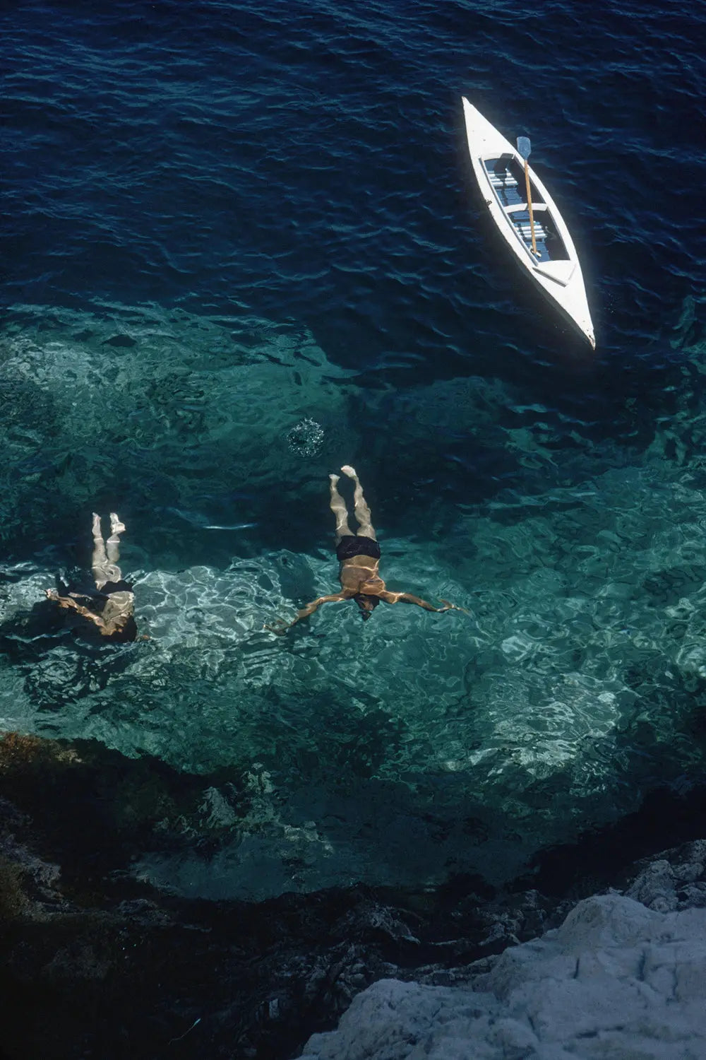 Aerial view of two people swimming in clear turquoise water near a small white boat on deep blue water, with rocky shoreline in the foreground. The Picturalist Slim Aarons Collection. Coastal Photography.