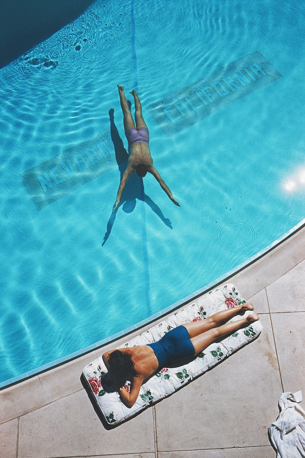 A top-down view of a swimming pool with a woman floating face down in the blue water and another woman lying on a floral-patterned pool float on the pool deck.