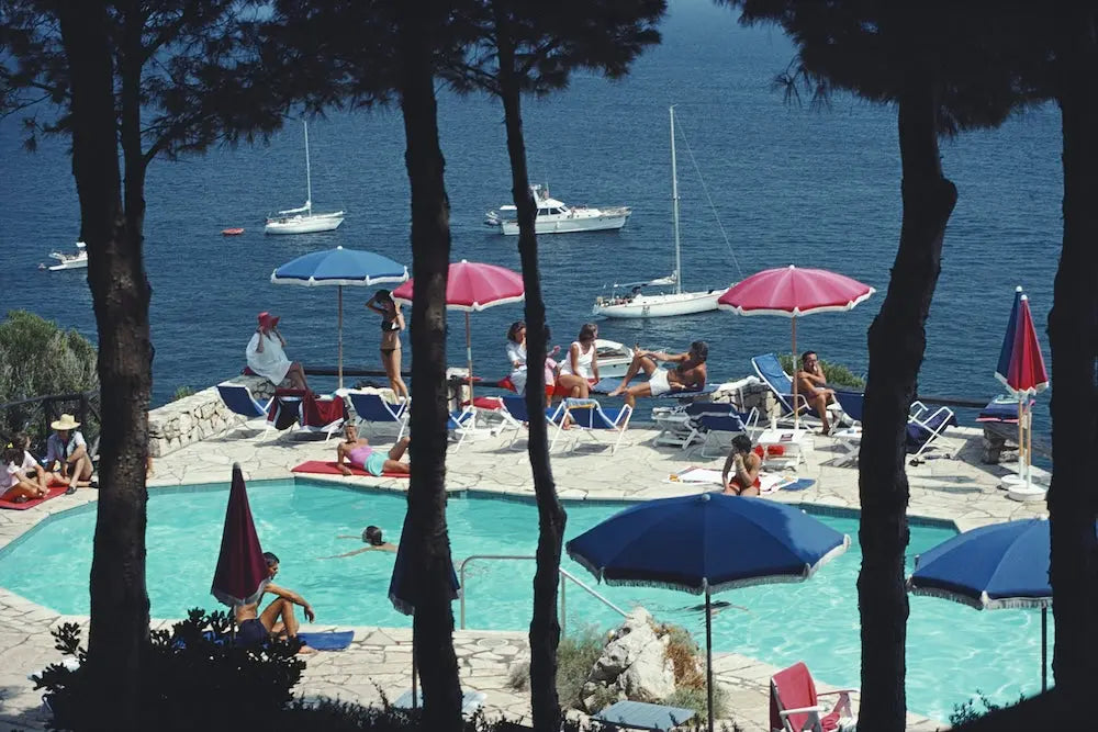 A scenic coastal resort scene with a swimming pool, lounge chairs, colorful umbrellas, people relaxing, sailboats on the blue sea, and dark tree trunks framing the foreground. The Picturalist Wall Art and Photography for Designed Interiors. Slim Aarons Collection, Poolside. Getty Images Gallery.