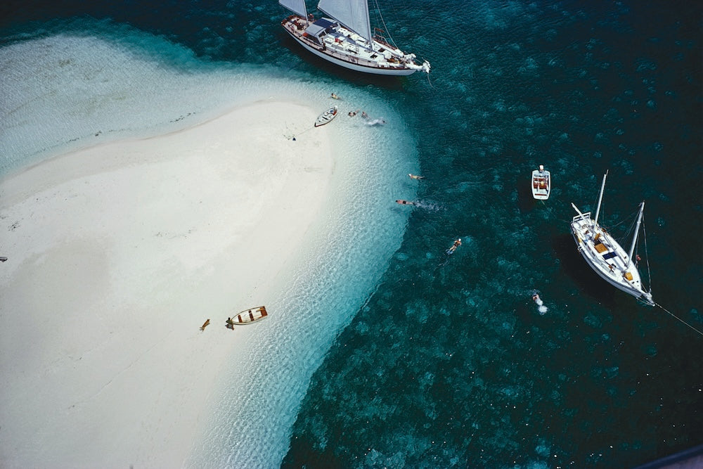 Aerial view of a tropical sandbar with clear turquoise water, several sailboats anchored nearby, and small swimmers in the ocean. The Picturalist Wall Art and Photography for Designed Interiors.