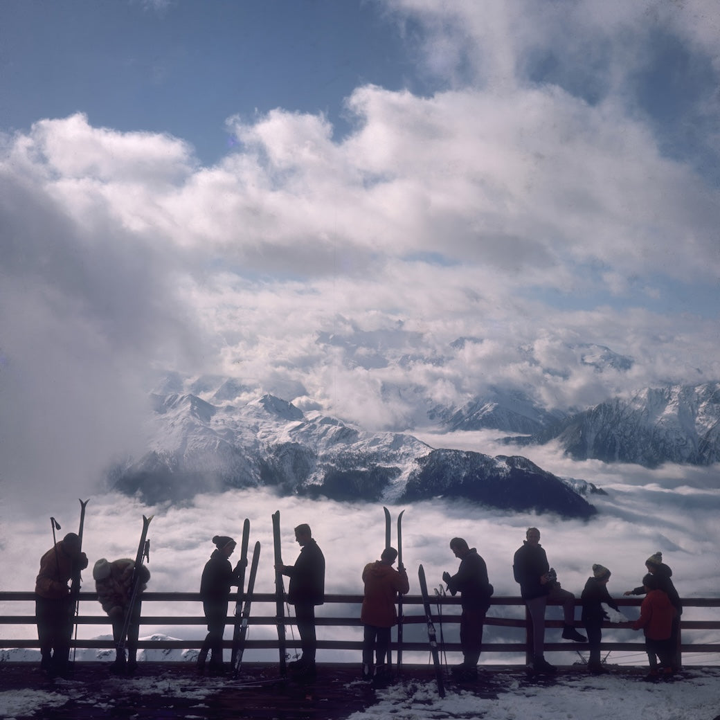 A scenic mountain landscape with snow-covered peaks rising above a sea of clouds, and a group of silhouetted people standing along a wooden railing holding skis in a high-altitude alpine setting.