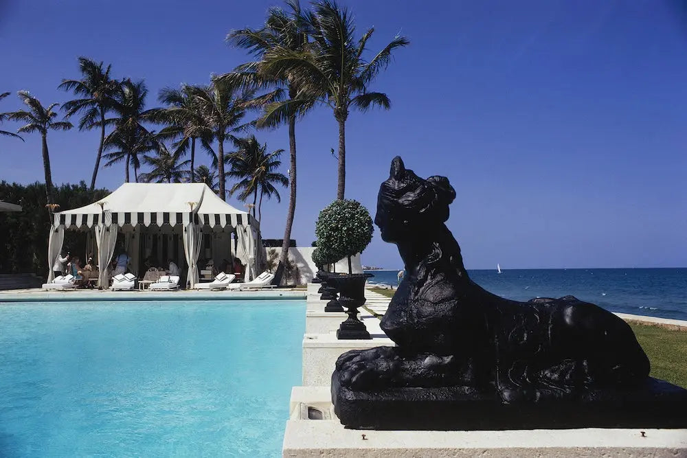 A luxurious resort scene with a bright blue swimming pool in the foreground, a black lion statue, white cabanas, tall palm trees, and the blue ocean under a clear sky. The Picturalist Slim Aarons Collection.