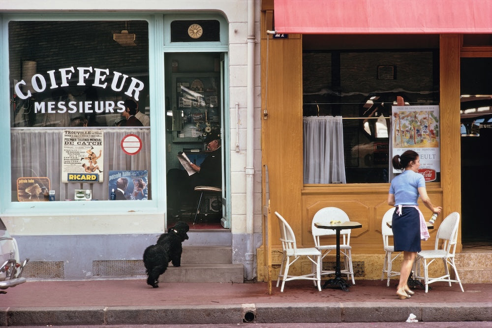 A street scene in front of a Parisian-style cafe with a turquoise window reading 'COIFFEUR MESSIEURS' and a wooden facade with a pink awning. White outdoor chairs and tables sit on the sidewalk next to a woman in a blue dress walking away, with a small black dog near the doorway.