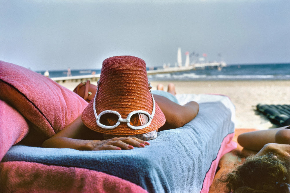 Person lying on a beach with a sun hat and sunglasses, ocean and skyline in the background. The Picturalist Slim Aarons Collection. Summer. Coastal Photography.