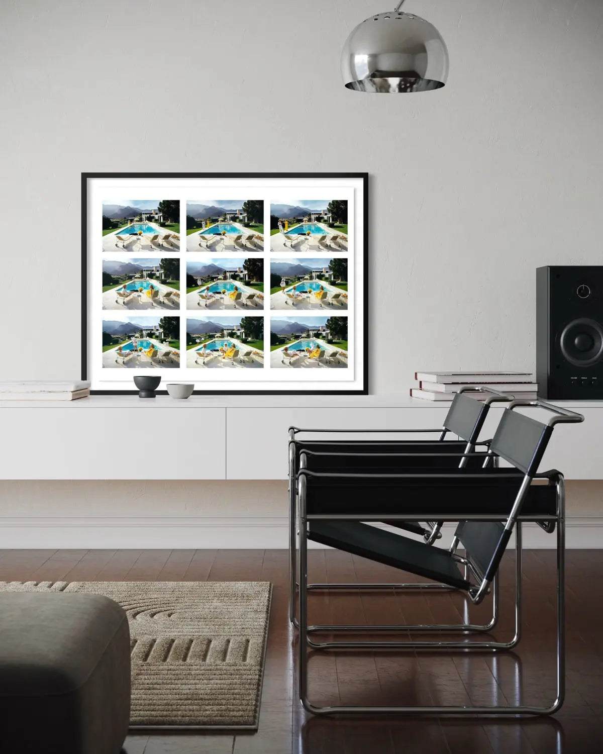 A framed art print featuring a 3x4 grid of aerial photographs of swimming pools, displayed on a white wall in a modern living room setting with two black chairs and a silver pendant light. The Picturalist Wall Art and Photography for Designed Interiors. Slim Aarons Collection, Poolside. Getty Images Gallery.
