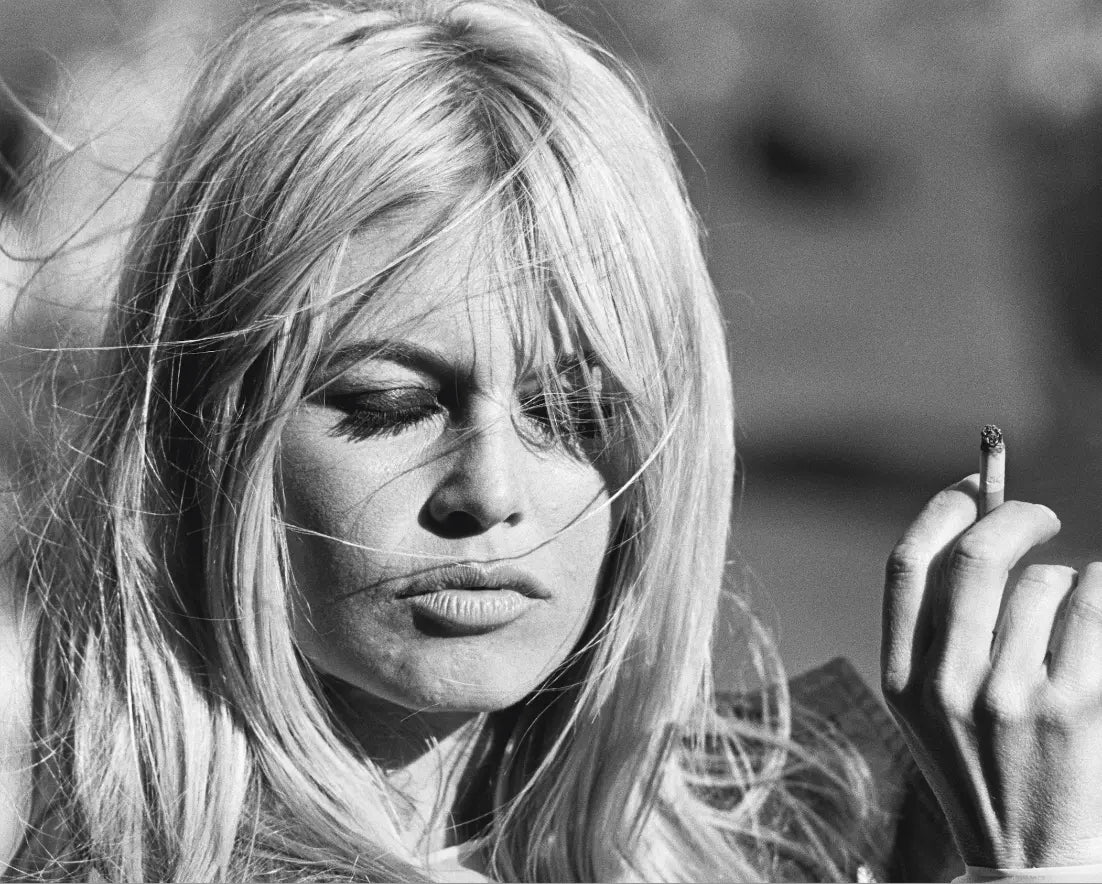 Black and white close-up portrait of actress Brigitte Bardot with long hair holding a cigarette, shown outdoors with a blurred background.