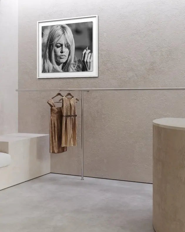 A framed black and white portrait print of actress Brigitte Bardot hangs on a textured beige wall above a minimalist clothing rack displaying two metallic bronze dresses in a modern, clean interior setting.