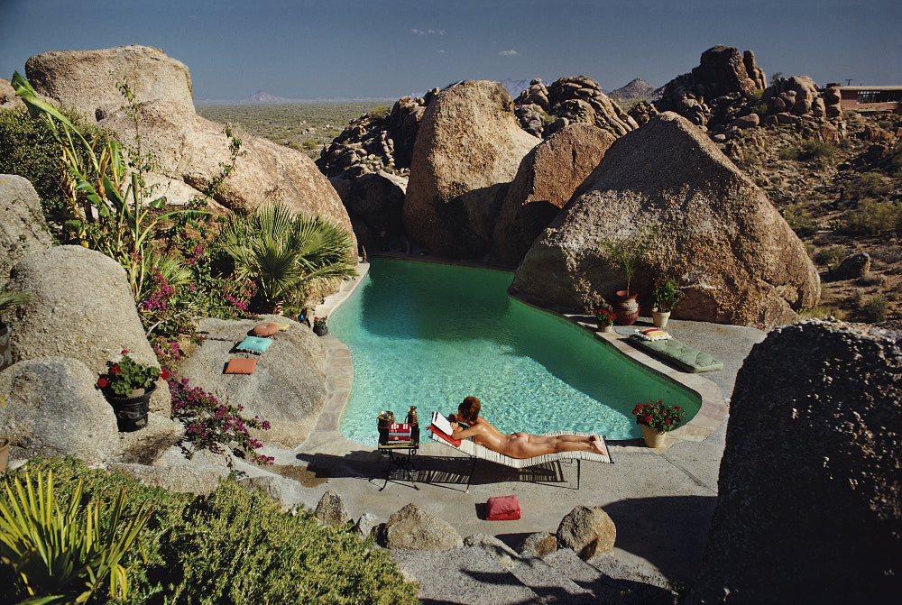 Person lounging by a pool surrounded by large rocks and desert landscape. The Picturalist Slim Aarons Collection.