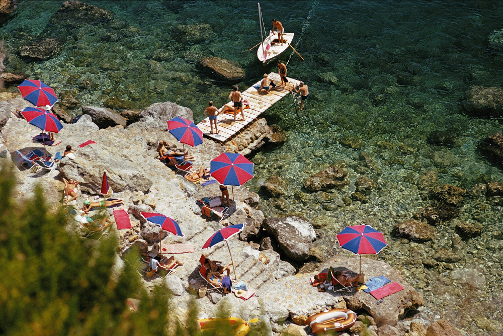 Il Pellicano inorto Ercole scene of people on a rocky beach with colorful umbrellas and a boat in the water. 88154880