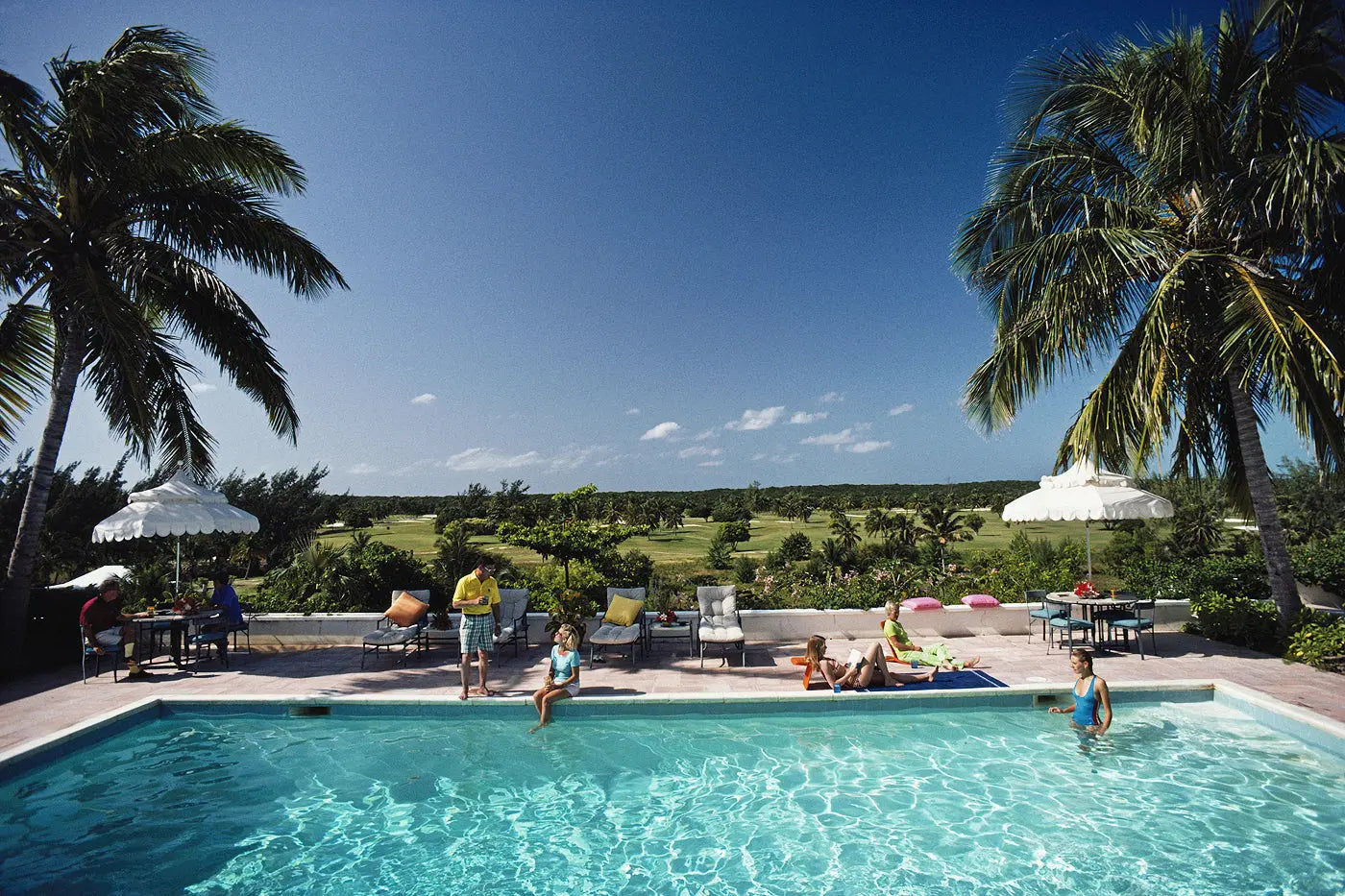 A resort-style swimming pool surrounded by lounge chairs, umbrellas, palm trees, and people relaxing outdoors under a clear blue sky.