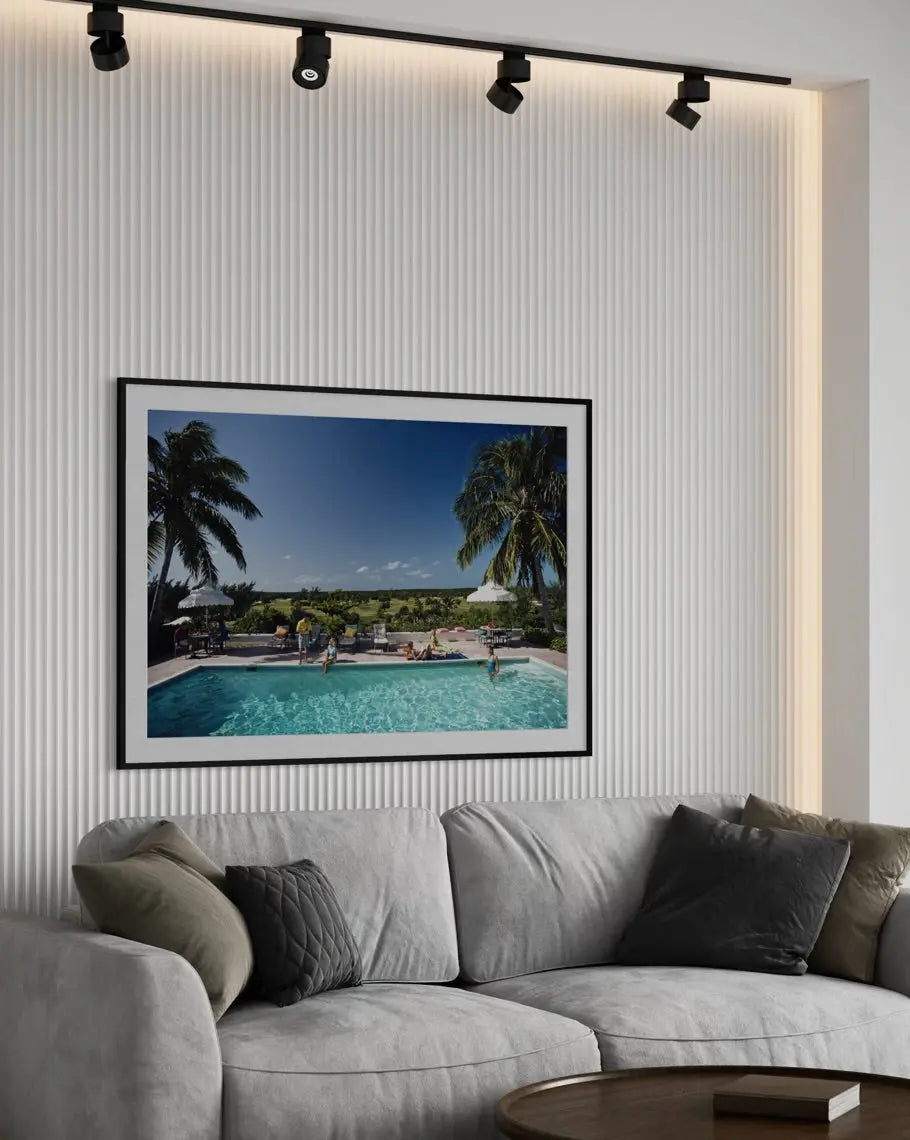 A framed art print depicting a tropical resort pool scene with palm trees and a blue sky, displayed on a ribbed white wall above a modern gray sofa in a living room setting.