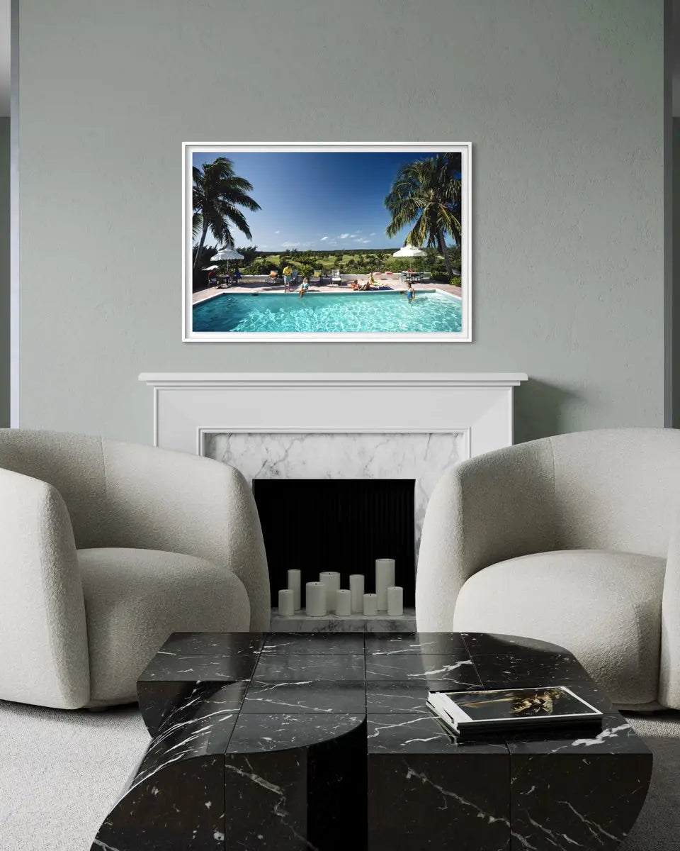 A framed art print depicting a tropical poolside scene with palm trees and a blue sky, displayed on a gray wall above a white fireplace mantel in a modern living room setting with two cream armchairs and a black marble coffee table.