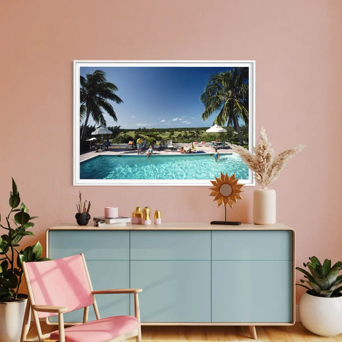 A framed wall art print depicting a tropical poolside scene with palm trees and a blue sky, displayed above a light blue console table in a styled room with a pink wall, chair, plants, and decorative objects.
