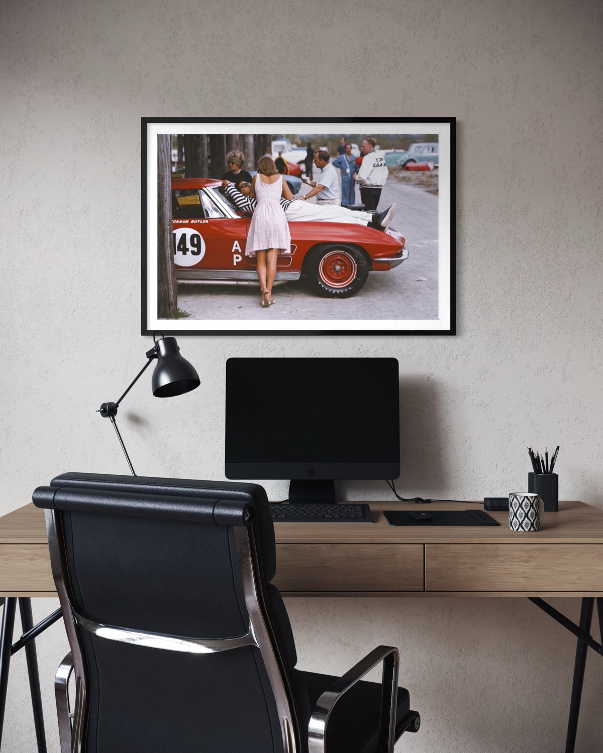 A framed art print depicting a vintage red race car scene with a woman in a white dress, displayed on a textured gray wall above a modern desk setup with a computer monitor, black office chair, and desk lamp.