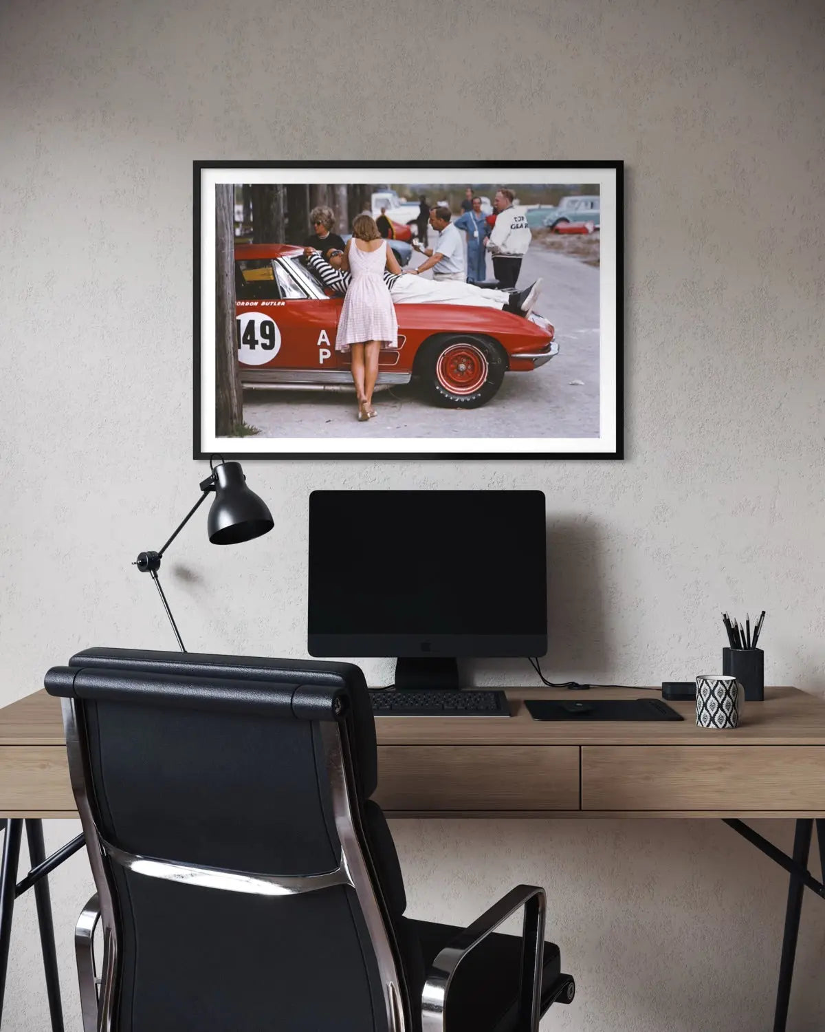 A framed art print depicting a vintage red race car scene with a woman in a white dress, displayed on a textured gray wall above a modern desk setup with a computer monitor, black office chair, and desk lamp.