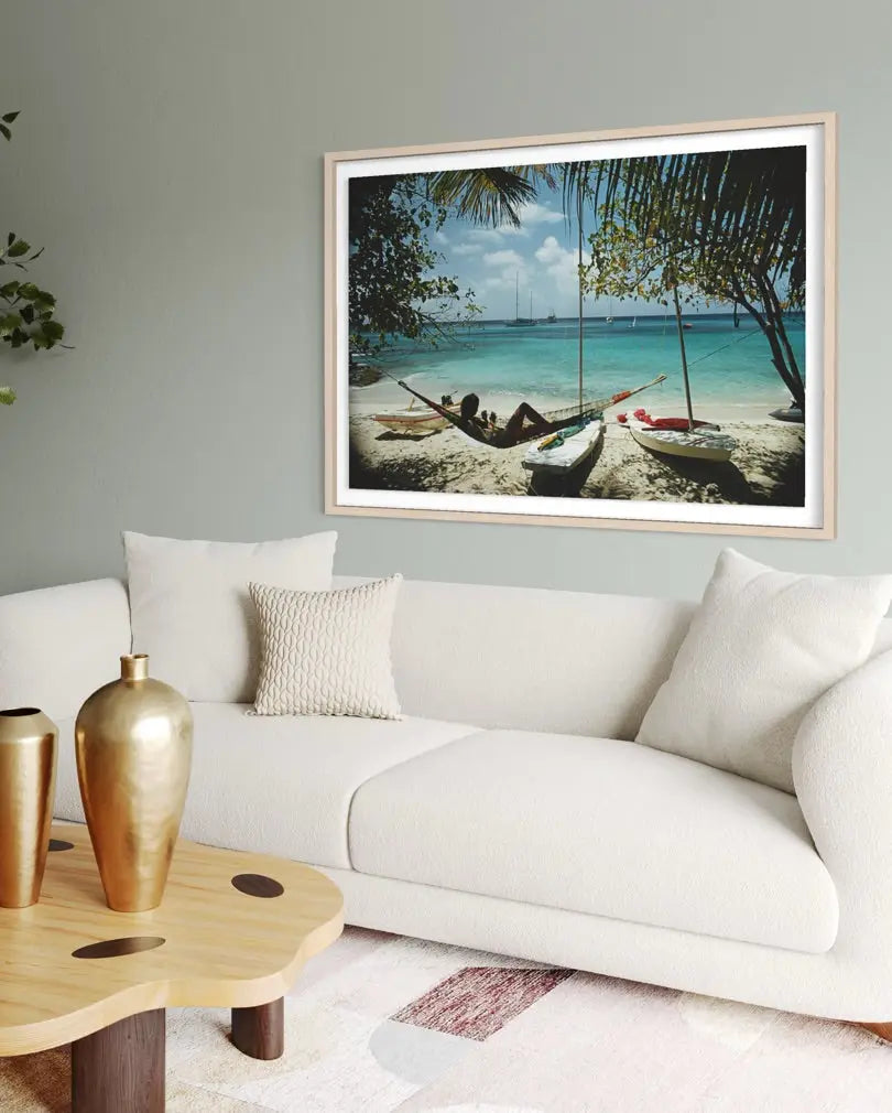 A framed beach scene print hangs on a light gray wall above a white sofa in a styled living room. The artwork shows a tropical shoreline with palm trees, blue water, and small boats, while the room background includes a white couch, pillows, a wooden coffee table, and gold vases.
