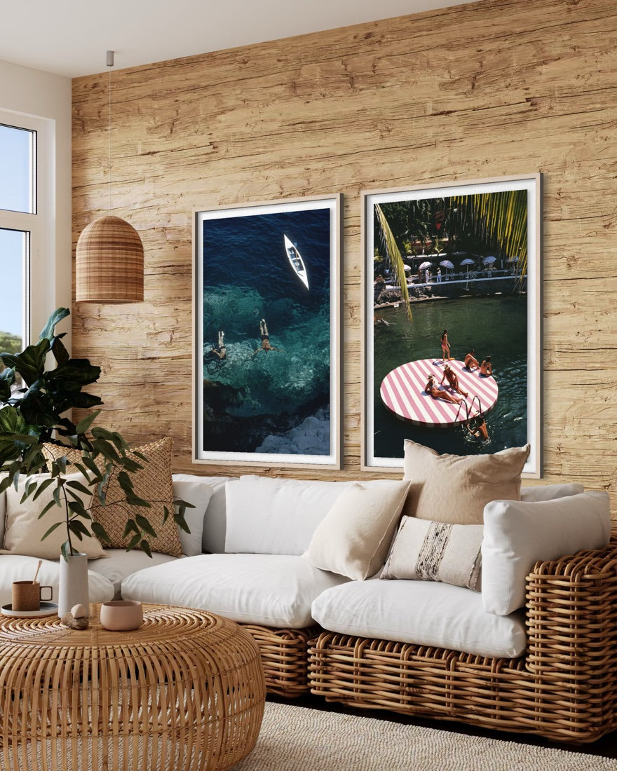 Two vertical framed art prints depicting tropical beach and pool scenes, displayed side-by-side on a rustic wood-paneled wall above a white and wicker sofa in a modern living room setting.