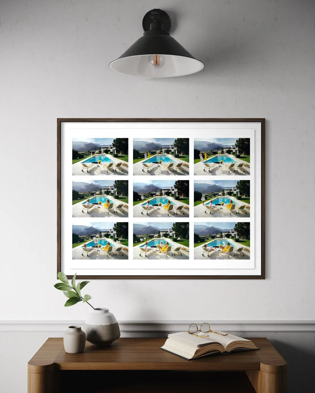 A framed art print featuring a 3x3 grid of nine photographs of a swimming pool and patio scene, hung on a light gray wall above a wooden console table with a vase, book, and glasses. The Picturalist Wall Art and Photography for Designed Interiors. Slim Aarons Collection, Poolside. Getty Images Gallery.