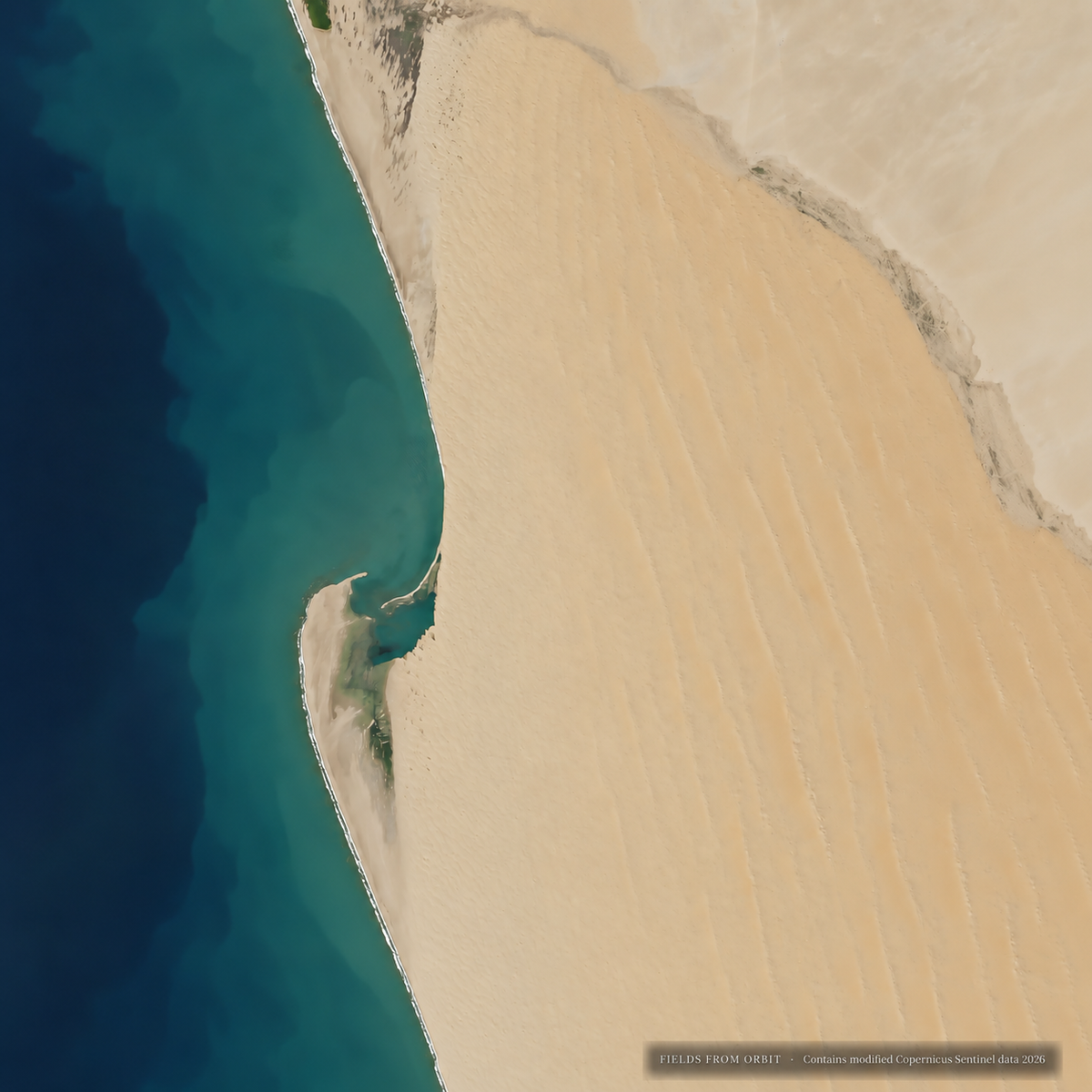 Aerial photograph of a narrow sandy shoreline curving into deep turquoise water, with pale beige sand and darker blue water filling the frame.