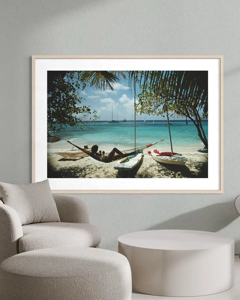 A framed beach art print hangs on a light gray wall above a modern beige armchair and round side table. The print shows a tropical beach scene with turquoise water, palm trees, and small boats on the sand.