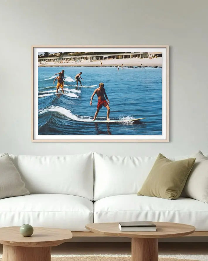 A framed photographic print of surfers riding waves in a sunny beach setting, displayed on a light gray wall above a modern white sofa with wooden coffee tables.