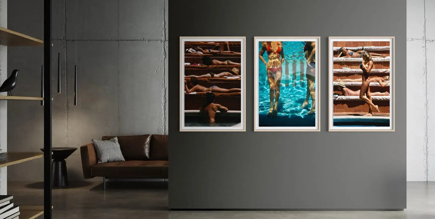 A three-panel framed wall art set hangs on a dark gray wall in a modern living room. The artwork depicts sunbathing figures on wooden decks and a swimmer in bright blue water, each panel in a white frame.