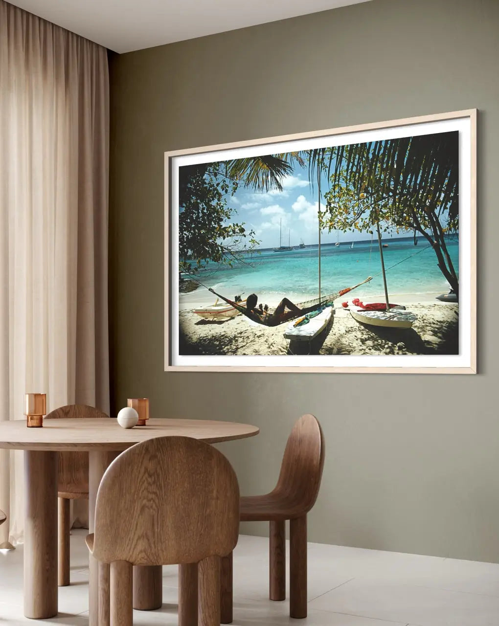 A framed tropical beach print hanging on a sage green wall in a dining room setting with a wooden table and chairs.