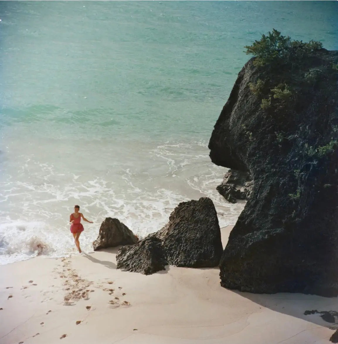 A vintage-style photograph of a beach scene with a person in a red swimsuit walking near the shoreline, large dark rocks, pale sand, and turquoise ocean water. The Picturalist Wall Art and Photography for Designed Interiors. Slim Aarons Collection, Coastal. Getty Images Gallery.