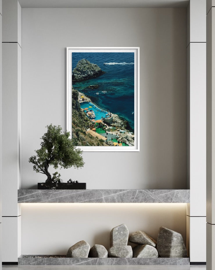 A framed vertical art print depicting a scenic coastal cove with turquoise blue water, rocky cliffs, and green hills, displayed on a light gray wall above a marble console with a small tree and stones.