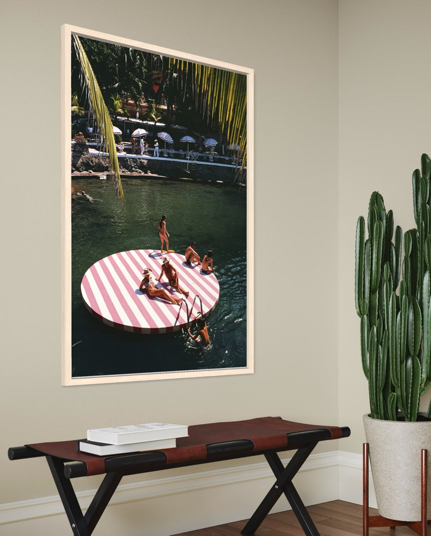 A framed vertical art print depicting a tropical poolside scene with a pink and white striped floating platform, displayed on a beige wall above a bench with a potted cactus nearby. The Picturalist Wall Art and Photography for Designed Interiors. Slim Aarons Collection, Coastal. Getty Images Gallery.