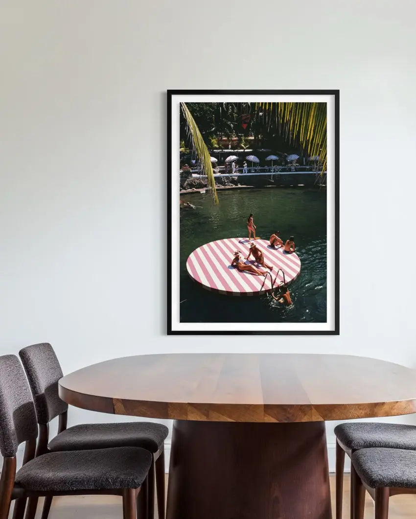 A framed vertical art print depicting a woman relaxing on a pink and white striped circular float in a tropical pool, displayed on a light gray wall above a wooden dining table and chairs. The Picturalist Wall Art and Photography for Designed Interiors. Slim Aarons Collection, Coastal. Getty Images Gallery.