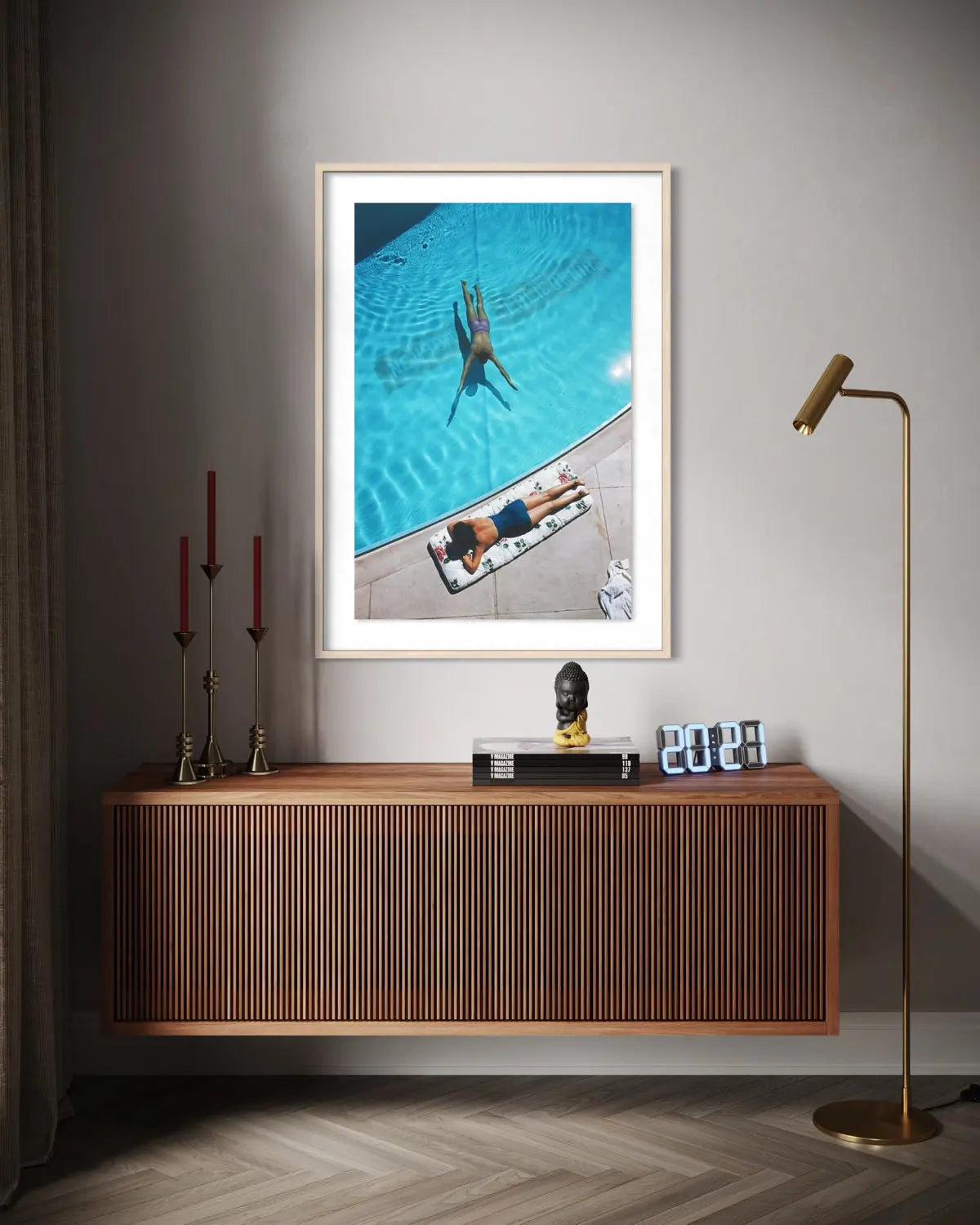 A framed art print depicting an aerial view of a person swimming in a blue pool next to another person sunbathing, styled on a gray wall above a wooden console table in a modern living room setting. The Picturalist Slim Aarons Collection.